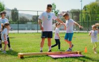 Kids guided by instructors at a park balance on a beam for fun and exercise.